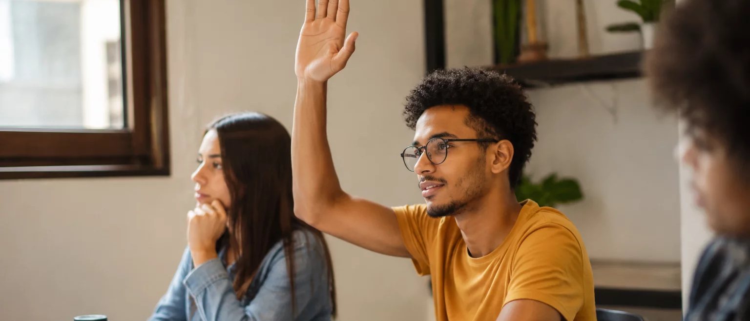 Participante com a mão erguida pedindo a vez, à mesa coberta de folhas escritas